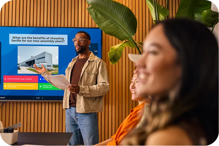 A male presenter leading an interactive business meeting, standing next to a large screen displaying a quiz question about the benefits of choosing Seville for a new assembly plant.