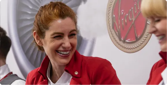 A candid photograph of two women in red uniforms laughing together. The woman in the center has red hair tied back and is smiling broadly. In the background, a circular "Virgin" logo is partially visible on a white wall next to a large turbine-like structure.