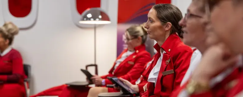 Cabin crew in red uniforms sit with tablets, listening during a training session.