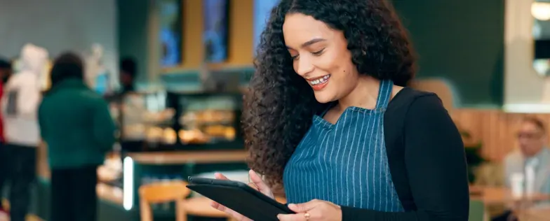 A smiling café employee in a striped apron uses a tablet at work.