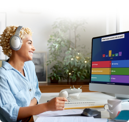 A woman with blonde curly hair wearing silver headphones smiles while looking at her computer. The monitor displays a live Kahoot! poll with colorful bar chart results.